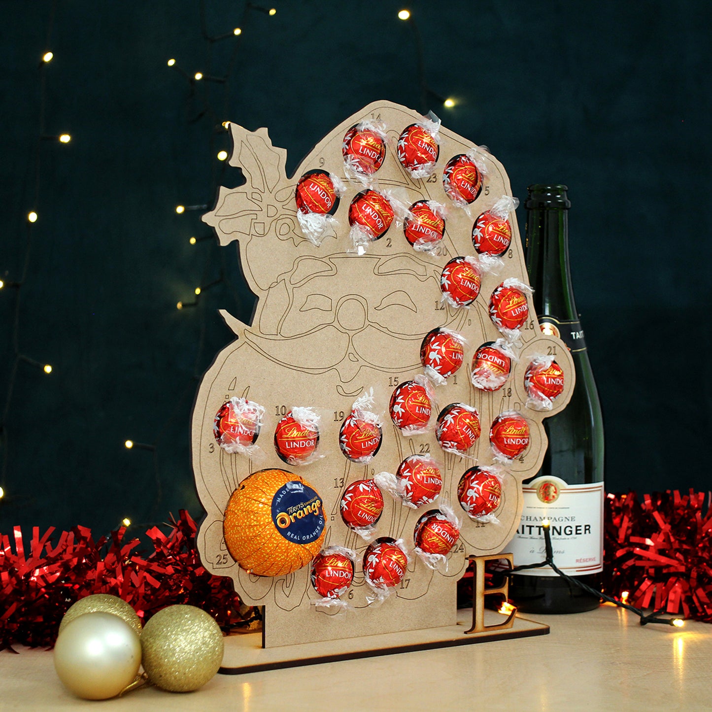 Santa face shaped MDF advent calendar filled with chocolate on table in front of black background with fairy lights