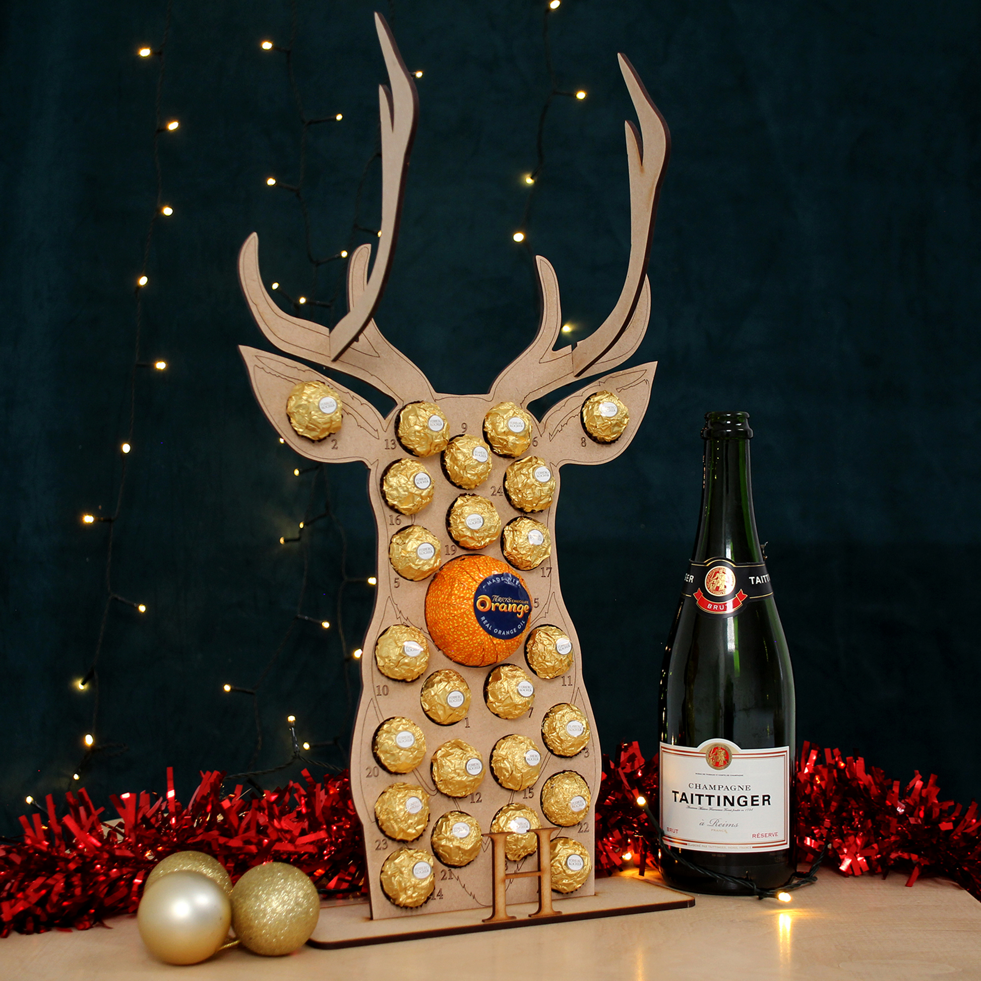 Reindeer shaped MDF advent calendar filled with chocolate on table in front of black background with fairy lights